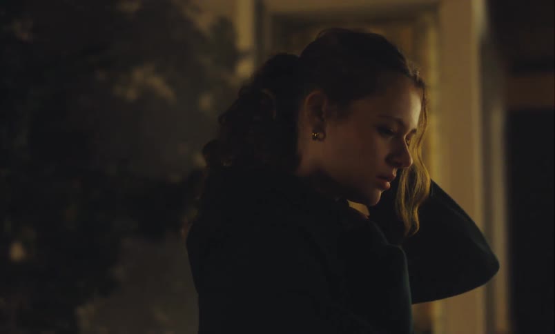 A sad and pensive young woman with curly hair stands alone outside at night, lost in thought
