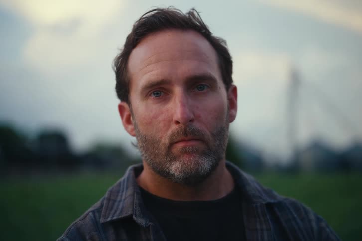 Portrait of a serious-looking middle-aged man with a beard coming into focus as he looks directly at the camera in a field at dusk
