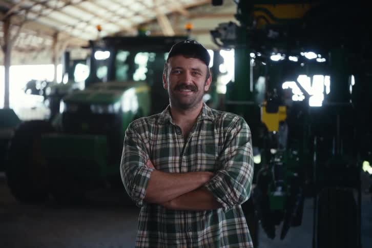 A proud farmer with a mustache and plaid shirt stands smiling with his arms crossed in a barn in front of his tractors