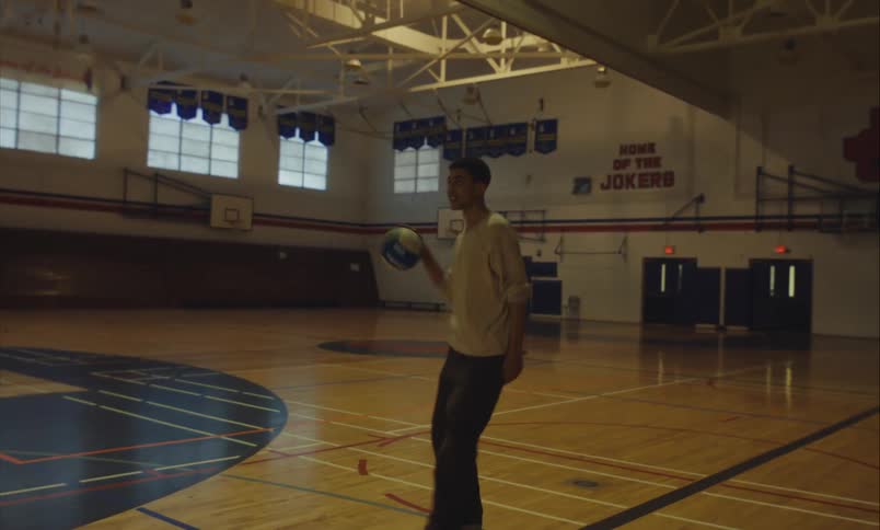 A young man practices volleyball alone in an empty high school gymnasium, playfully shooting baskets and waving