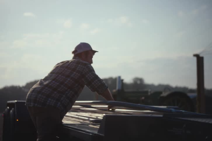 A hardworking man in a plaid shirt secures equipment onto a flatbed trailer with ratchet straps in a rural field at sunrise