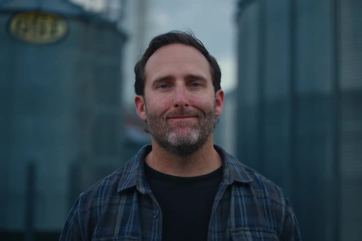 A proud farmer in a plaid shirt smiles confidently and crosses his arms in front of large grain silos