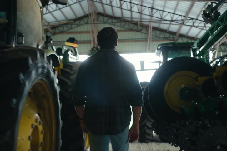 A farmer in a plaid shirt and jeans walks slowly through a large machine shed filled with modern tractors and agricultural equipment