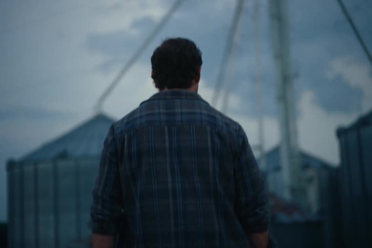 A farmer in a plaid shirt and boots walks in slow motion towards grain silos at dusk