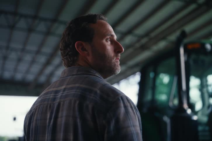 A serious farmer wearing a plaid shirt stands thoughtfully inside a large barn next to his tractor