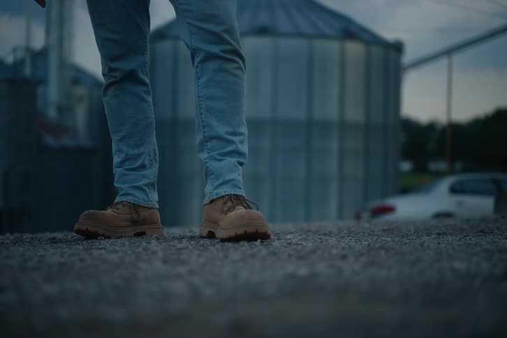 A low angle static shot of a person's boots and jeans standing on a gravel road in front of a large farm silo at dusk