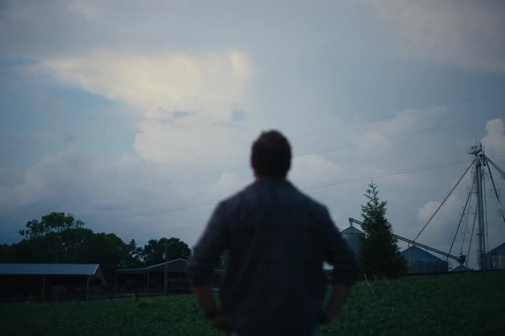 A farmer stands with hands on his hips, thoughtfully observing his fields and silos as evening approaches
