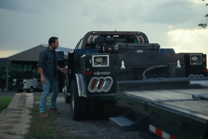 A man in a flannel shirt and jeans gets out of his work truck and grabs a wrench from a toolbox on the flatbed