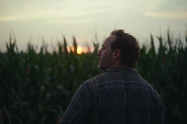 A contemplative farmer in a plaid shirt looks out over his bountiful cornfield at sunset