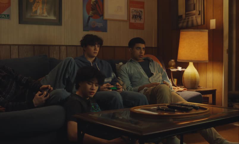 A diverse group of teenage boys hang out in a dimly lit living room playing video games and using their smartphones