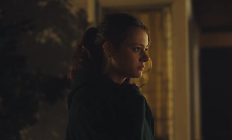 A young woman stands alone in a dimly lit room, her sad expression hinting at deep thought and loneliness
