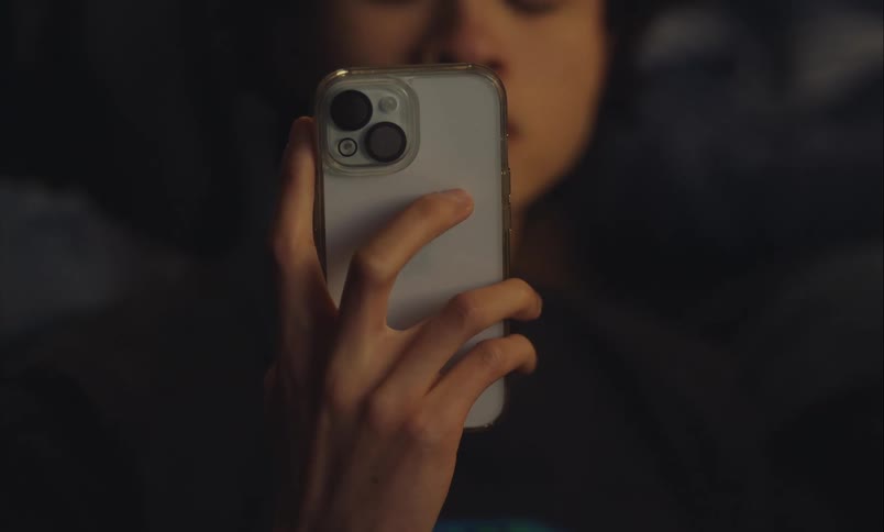 A close-up shot of a person's hand holding a smartphone while they are lying down in a dark room