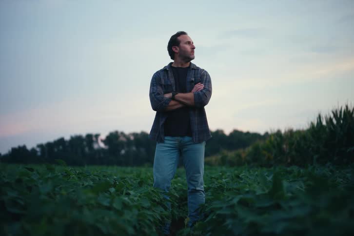A concerned farmer stands in his crop field at dusk, crossing his arms as he surveys his land