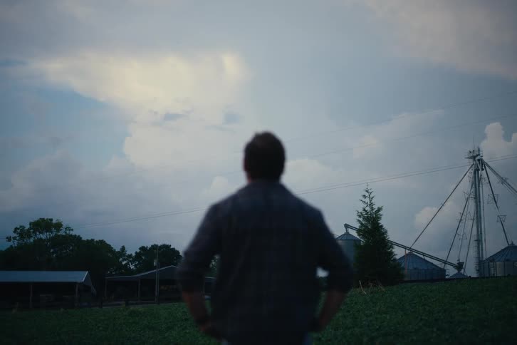 A farmer in soft focus surveys his farm and grain silos under a vast, cloudy sky at dusk
