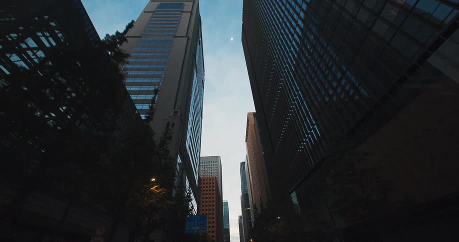 A cinematic forward tracking shot through a modern city street lined with tall glass skyscrapers at twilight