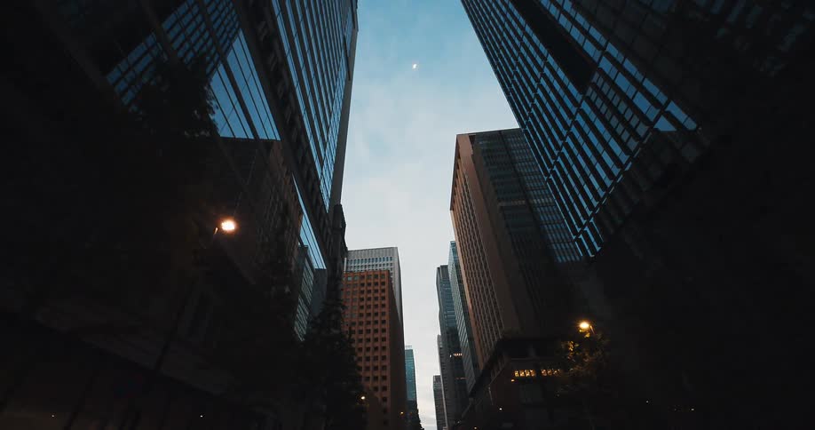 A slow, low-angle tracking shot moves through a modern city street, looking up at towering skyscrapers against the dusky sky
