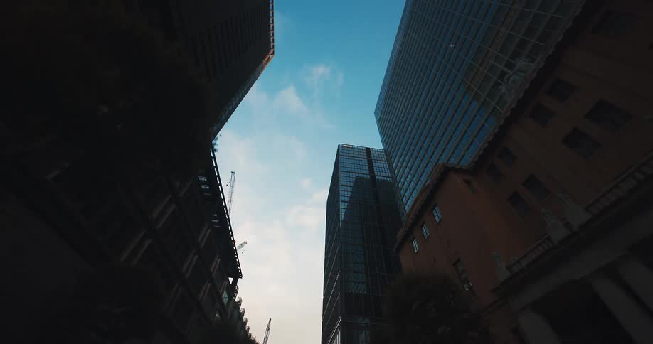 A slow, backward dolly shot looking up from a city street at towering modern skyscrapers silhouetted against the sky at dusk