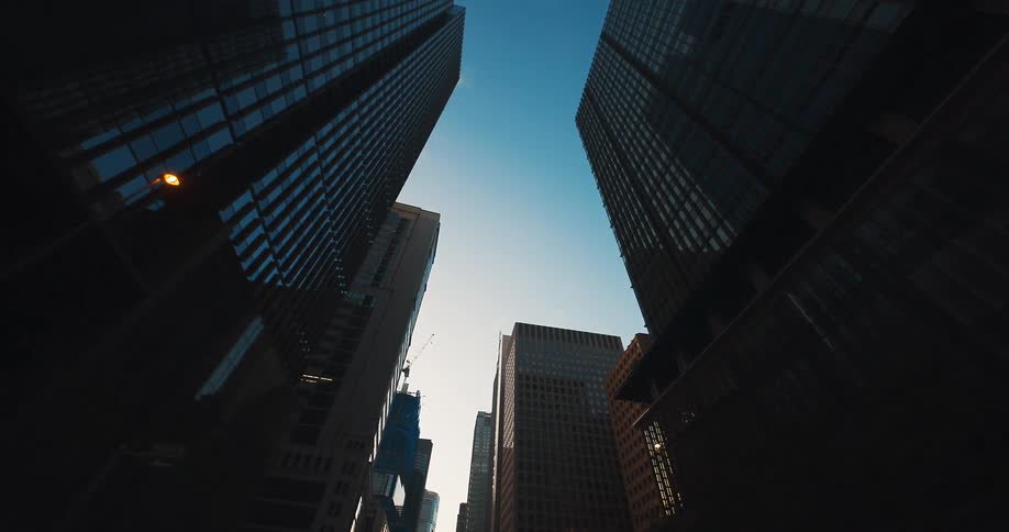 A dramatic, low-angle dolly shot moves backward between silhouetted skyscrapers, revealing modern architecture against a clear blue sky