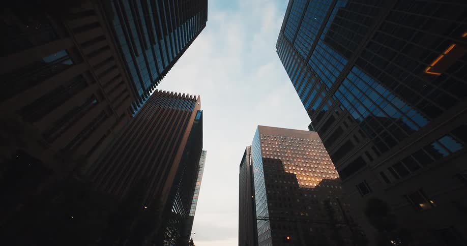 A slow forward-moving, low-angle shot captures the towering modern skyscrapers of a city financial district against the sky