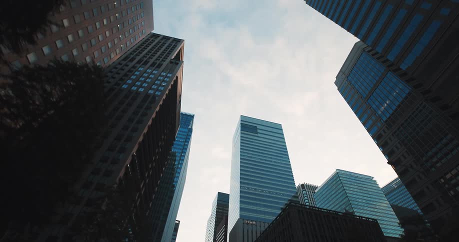 A slow moving, low-angle shot looking up at towering glass skyscrapers in a modern financial district against a lightly clouded sky