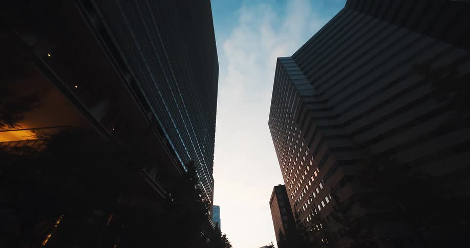 A low-angle reverse dolly shot glides between towering silhouetted skyscrapers in a modern city at dawn