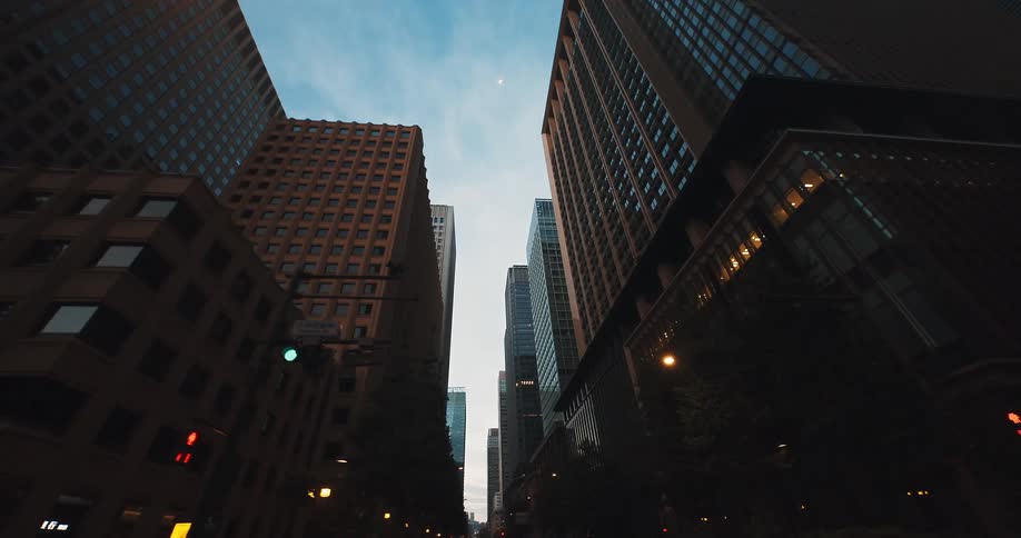 A slow, low-angle tracking shot moves through a canyon of modern skyscrapers in a quiet city street at twilight