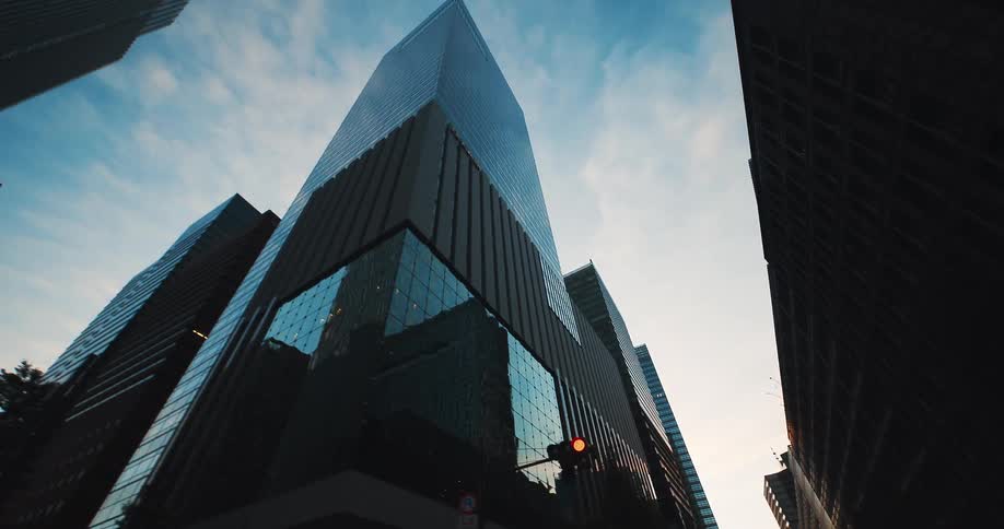 A low-angle tracking shot looks up at towering modern glass skyscrapers against the sky at dusk