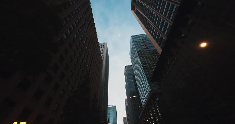 A slow, low-angle tracking shot looks up at towering city skyscrapers against the early evening sky as the camera moves down the street