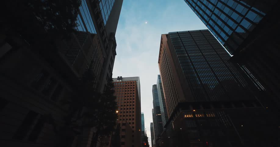 A slow dolly shot moves backwards along a city street, looking up at imposing skyscrapers and the moon at dusk