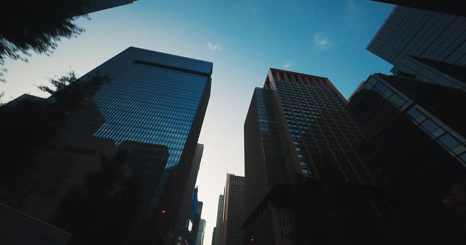 A slow, low-angle tracking shot moves through a modern city canyon of towering glass skyscrapers against a clear blue sky