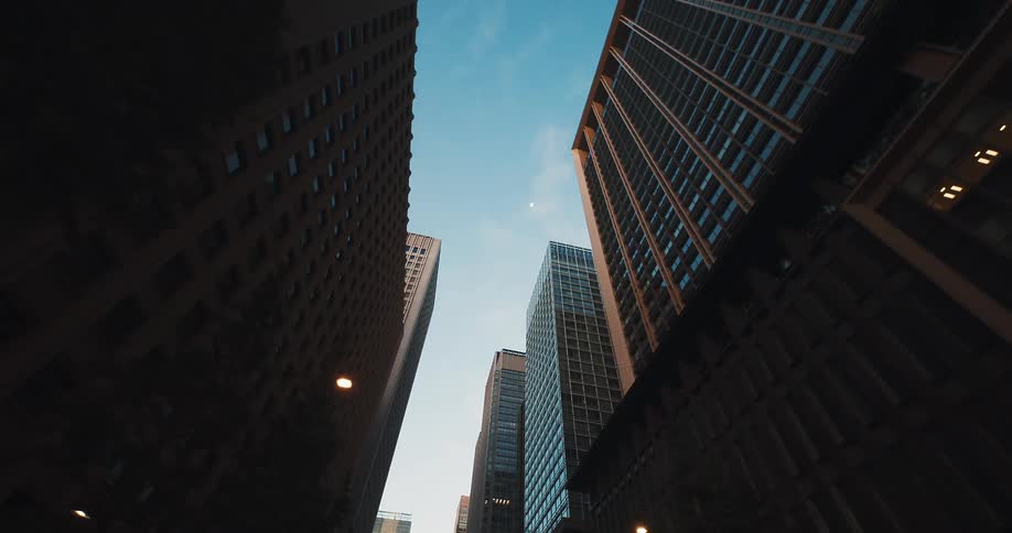 A slow tracking shot moves backward along a city street, looking up at towering skyscrapers against a clear blue evening sky