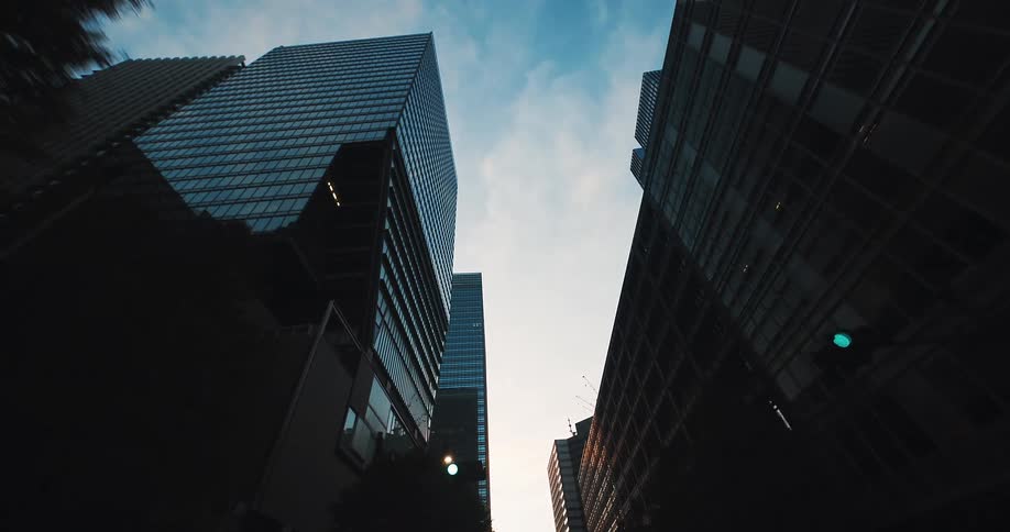A slow, low-angle tracking shot looks up at the towering skyscrapers of a modern downtown financial district silhouetted against the sky