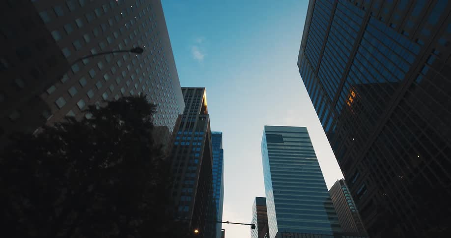 A dynamic, low-angle tracking shot moves through a modern city canyon of towering glass skyscrapers against a clear blue sky