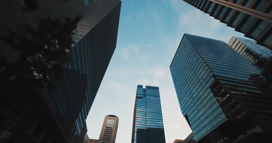 A smooth, low-angle tracking shot moves through a modern city, looking up at towering glass skyscrapers against a blue sky with a faint moon