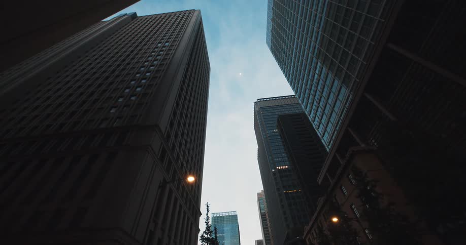 A low-angle tracking shot slowly pulls back from towering skyscrapers silhouetted against a beautiful dusk sky with a crescent moon