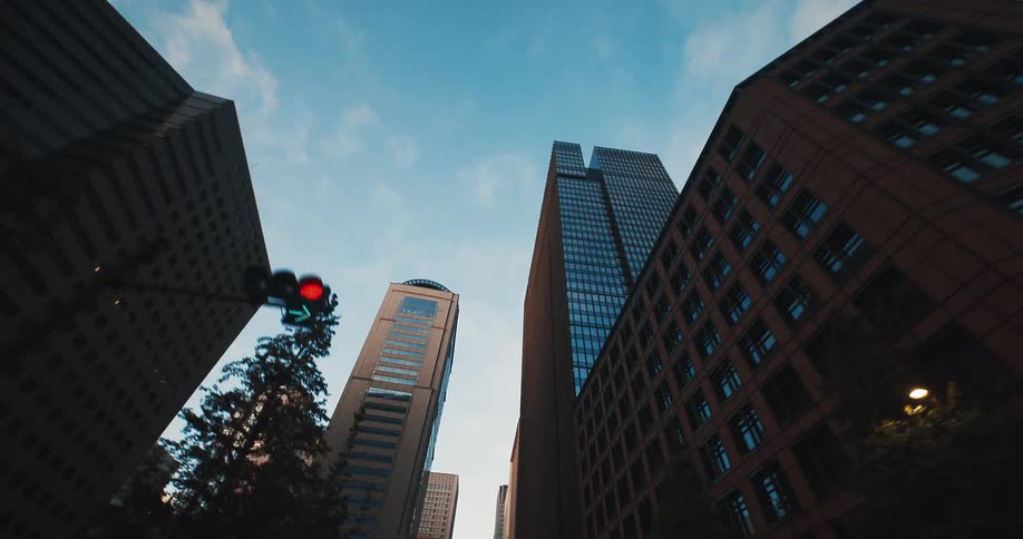 A smooth backward tracking shot looks up at towering modern skyscrapers in a bustling city against a blue sky