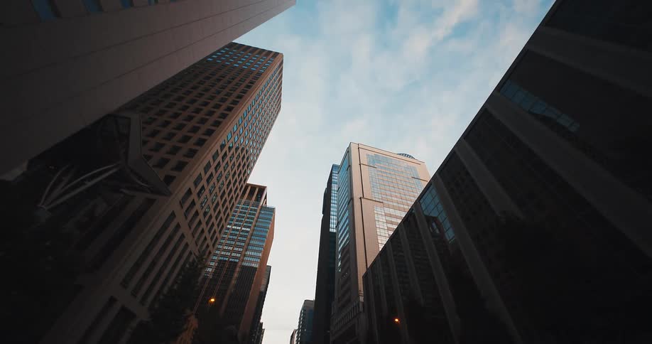 A low-angle tracking shot moves backward along a city street, looking up at modern skyscrapers against a partly cloudy sky