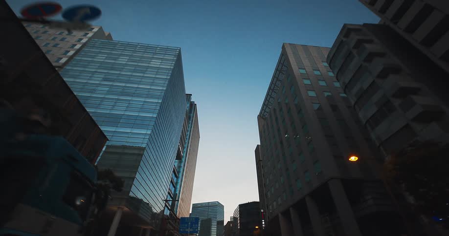 A serene low-angle dolly shot travels backward through a modern city street, revealing towering skyscrapers against the clear twilight sky
