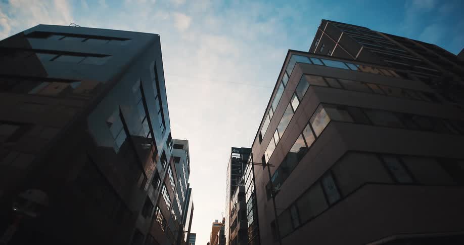 A slow, low-angle tracking shot moves backward through a narrow city street, looking up at the silhouetted facades of tall modern buildings against a bright sky