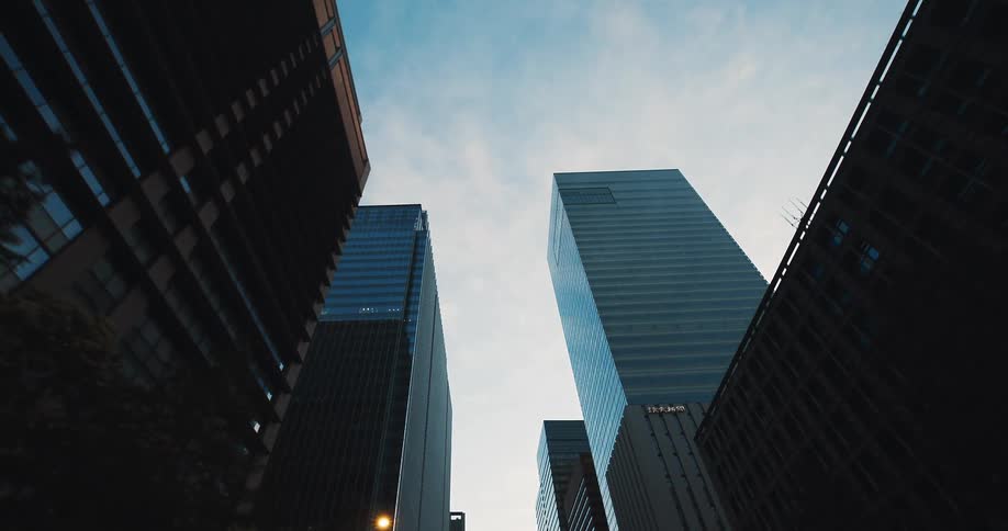 A slow, low-angle tracking shot through a canyon of modern glass skyscrapers reaching towards the blue sky