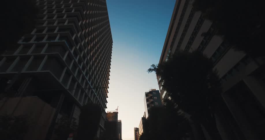 A smooth, low-angle forward tracking shot through a city canyon of silhouetted skyscrapers against a clear morning sky