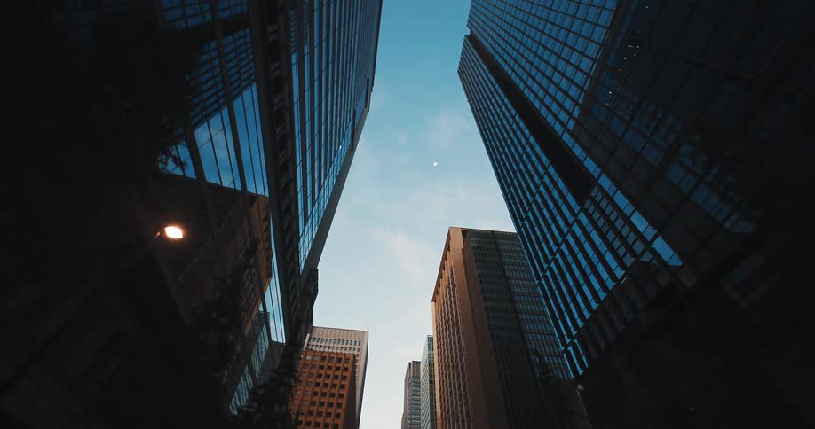 A low-angle perspective moves slowly through a metropolitan business district, looking up at towering glass skyscrapers against a clear blue sky