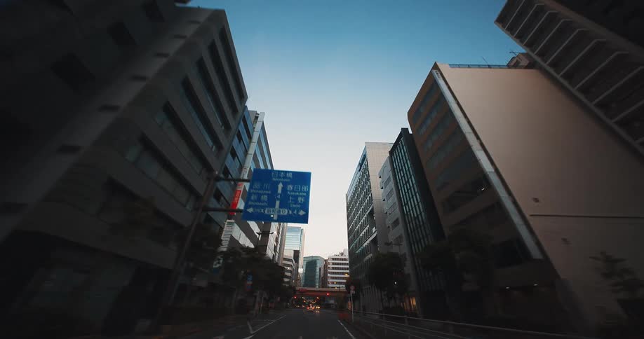 A serene point-of-view drive through the quiet streets of a modern Japanese city at dawn, looking up at the towering skyscrapers against a clear blue sky