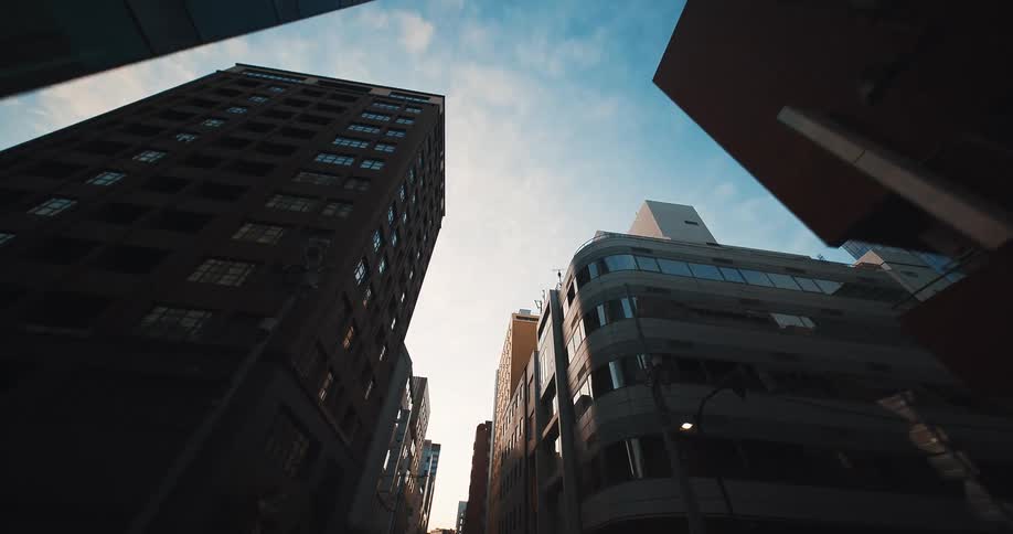 A smooth tracking shot glides down a narrow city street, with the camera pointed up at the towering buildings and the sky at dawn