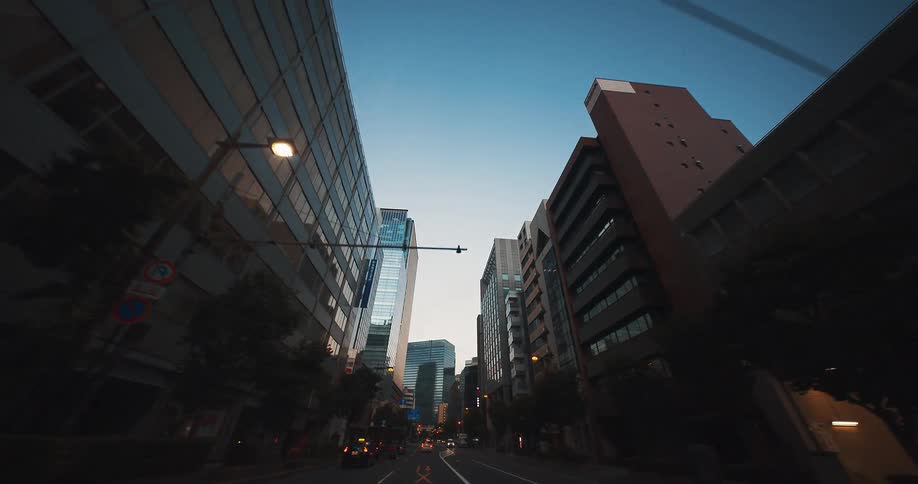 A slow, low-angle point-of-view drive through a canyon of modern city buildings at dawn in Japan