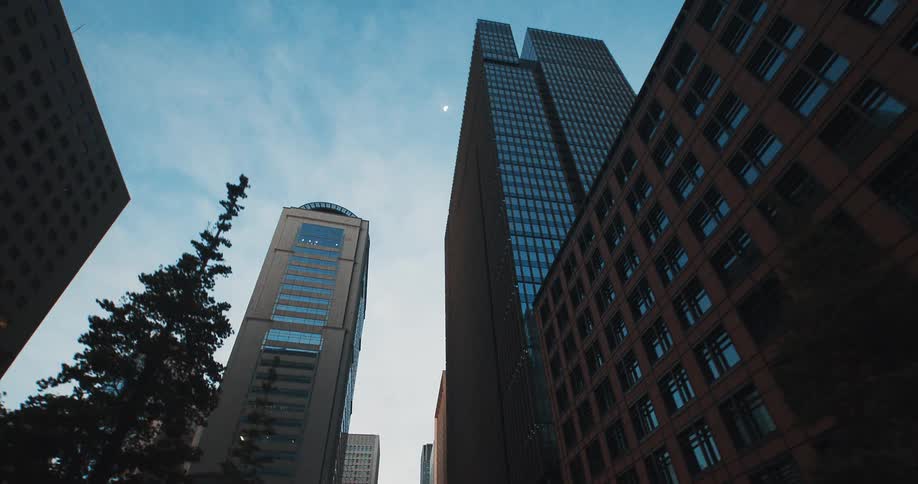 A smooth reverse tracking shot looks up at towering glass and steel skyscrapers in a modern financial district at dusk