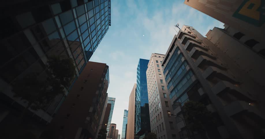 A slow, backward-moving, low-angle view reveals towering modern skyscrapers against a bright blue sky in a bustling city