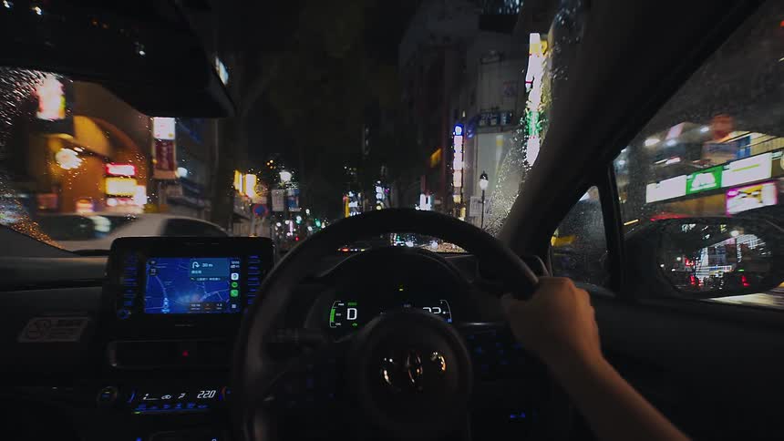 A first-person view from inside a car driving through the neon-lit streets of a Japanese city on a rainy night