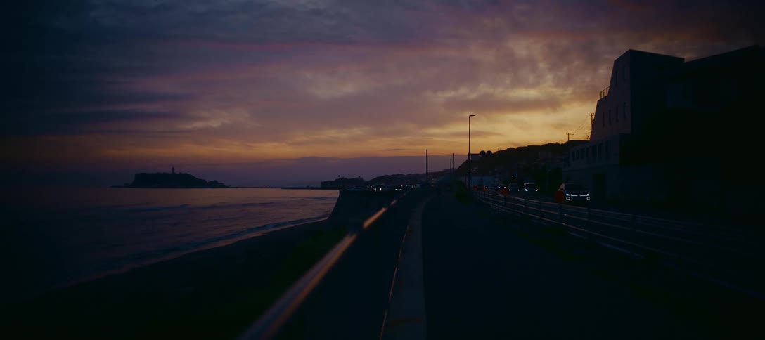 Traffic moves along a coastal highway at twilight as the sky glows with the colors of sunset over the ocean and a distant island