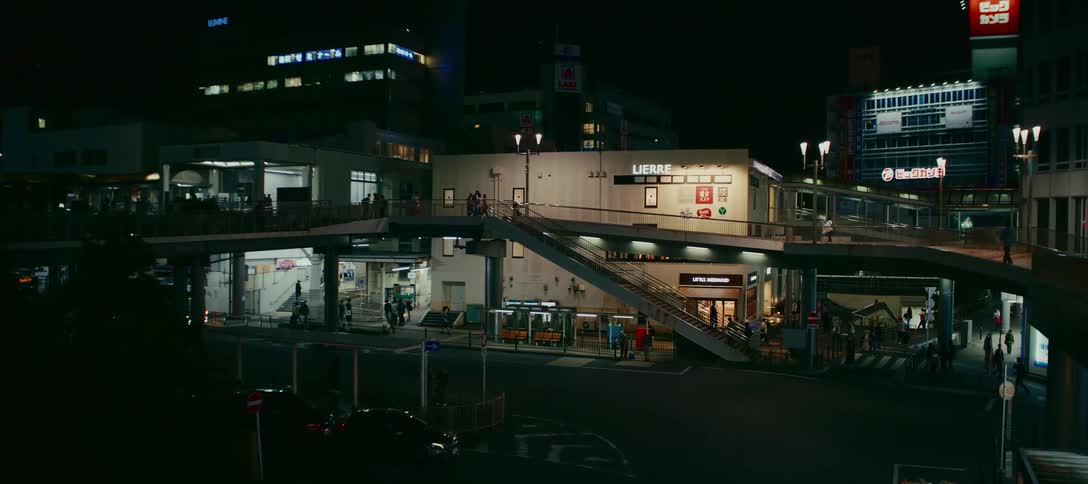 A bustling modern Japanese transit station and shopping complex illuminates the city street at night